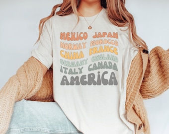 May include: A white t-shirt with a graphic design of various countries' names in a retro font. The countries listed are Mexico, Japan, Norway, Morocco, China, France, Germany, England, Italy, Canada, and America.