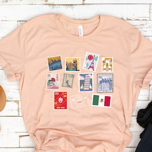 Op de afbeelding: Een lichtroze T-shirt met een afbeelding van kleurrijke postzegels met daarop bezienswaardigheden van over de hele wereld. De postzegels bevatten afbeeldingen van de Eiffeltoren, de Grote Muur van China, het Vrijheidsbeeld en meer.