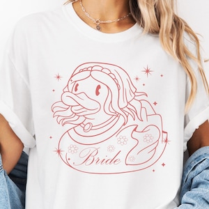 Könnte beinhalten: Weißes T-Shirt mit einer rosa Umrandung eines Entchens, das eine Krone trägt, und dem Wort "Bride" darunter. Das Entchen ist von kleinen Sternen umgeben.