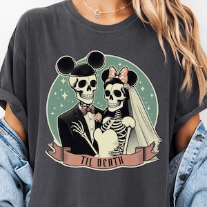 Puede incluir: Una camiseta gris oscuro con un gráfico de una pareja de esqueletos vestidos como Mickey y Minnie Mouse. El texto "Til Death" está impreso debajo del gráfico.