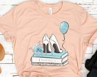 Puede incluir: Una camiseta de color melocotón con un diseño gráfico de un par de tacones altos blancos sobre una pila de libros con la palabra "Cenicienta" en la parte delantera del libro superior. Un globo azul con purpurina flota sobre los zapatos.