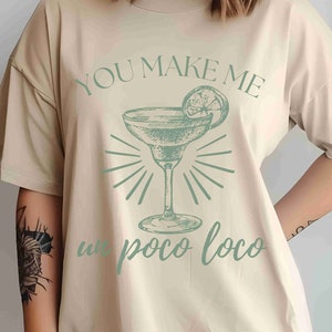 May include: Beige t-shirt featuring a margarita graphic and the text "You Make Me Un Poco Loco" in light green. The graphic shows a margarita glass with a lime slice. The shirt has short sleeves and a relaxed fit.
