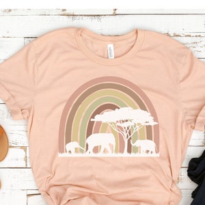 Op de afbeelding: Een perzikkleurig T-shirt met een regenboog en drie olifanten die onder een boom lopen. De regenboog is in tinten bruin, groen en geel.