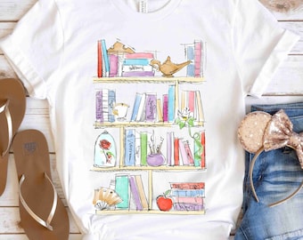 May include: White t-shirt with a graphic of a bookshelf filled with books and Disney characters. The bookshelf has a rose, a teapot, a genie lamp, and a book with an apple on it.