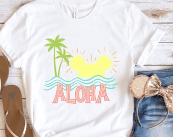 May include: White t-shirt featuring a graphic of a yellow sun with a Mickey Mouse silhouette in the centre, palm trees, and the word "ALOHA" in pink and blue lettering. The shirt is on a wooden surface with a pair of brown flip-flops and a pair of denim shorts with a gold glitter headband.
