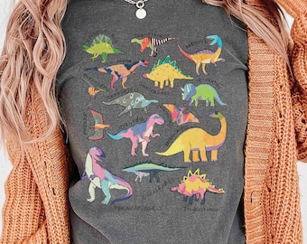May include: A dark gray t-shirt with a colorful dinosaur graphic print. The print features various dinosaurs with their names written underneath. The shirt is being worn by a person with long blonde hair.