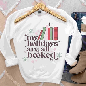 Op de afbeelding: Witte trui met een afbeelding van een stapel boeken en de tekst "my holidays are all booked". De afbeelding is in roze, groen, rood en wit. De trui ligt op een wit pluche oppervlak met een blauwe spijkerbroek, een bruine leren laars en een koffiebeker.
