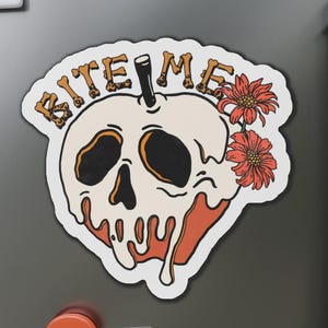 Puede incluir: Una pegatina con un diseño de calavera y las palabras "BITE ME" en letras con forma de hueso. La calavera es de color crema con cuencas de ojos negras y un diseño naranja goteando. Dos flores naranjas están en el lado derecho.