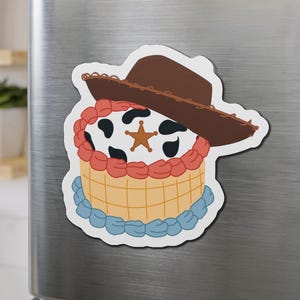 May include: A cartoon magnet depicting a cake with a cowboy hat. The cake has a cow-print top, red frosting, and a light brown base. A gold star-shaped badge is in the center of the cake. The magnet is on a silver surface.