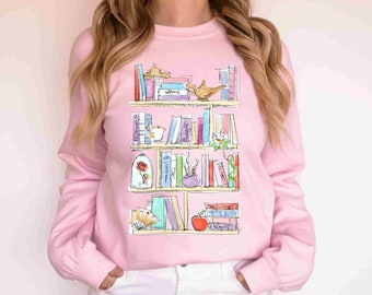 May include: A pink sweatshirt with a graphic of a bookshelf filled with books, a lamp, a rose, a teacup, and an apple. The books have titles like "Beauty and the Beast" and "Aladdin".