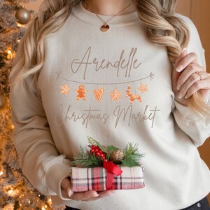 May include: A beige sweatshirt with the text "Arendelle Christmas Market" and a string of gingerbread cookies, stars, and hearts.