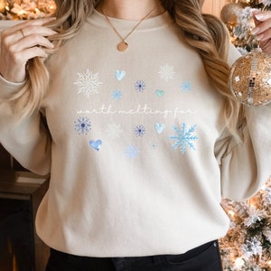 Puede incluir: Una sudadera beige con un dise&ntilde;o de invierno con copos de nieve y corazones. El texto "worth melting for" est&aacute; impreso en la parte delantera de la sudadera.