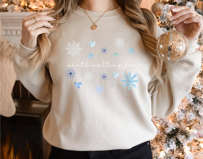 Puede incluir: Una sudadera beige con un dise&ntilde;o de invierno con copos de nieve y corazones. El texto "worth melting for" est&aacute; impreso en la parte delantera de la sudadera.