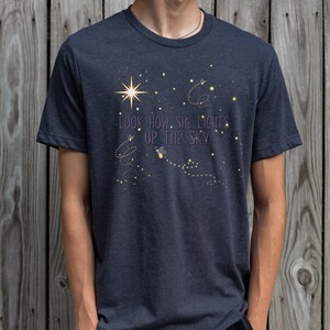 May include: A dark heather grey t-shirt with a graphic of fireflies lighting up the sky. The text "LOOK HOW SHE LIGHTS UP THE SKY" is printed on the shirt.