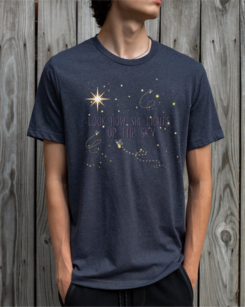 May include: A dark heather grey t-shirt with a graphic of fireflies lighting up the sky. The text "LOOK HOW SHE LIGHTS UP THE SKY" is printed on the shirt.