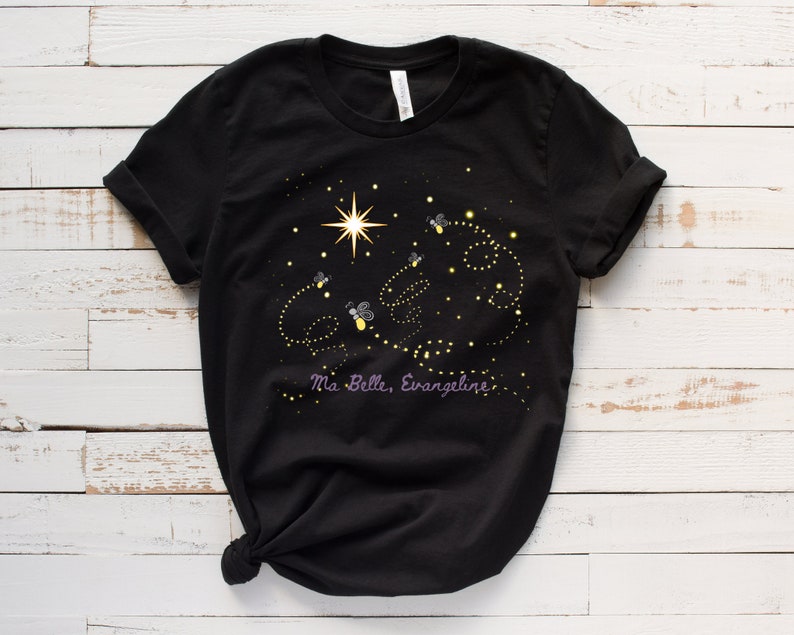 Puede incluir: Camiseta negra con un dise&ntilde;o de luci&eacute;rnagas y una estrella en un cielo nocturno. El texto "Ma Belle, Evangeline" est&aacute; impreso en morado debajo del dise&ntilde;o.