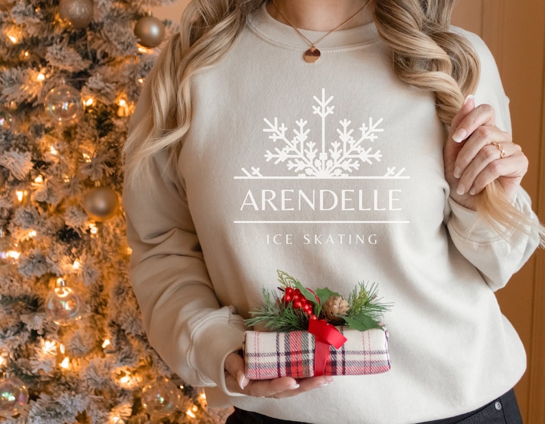 K&ouml;nnte beinhalten: Ein beiger Pullover mit einem wei&szlig;en Schneeflocken-Design und dem Text "Arendelle Ice Skating". Die Person h&auml;lt eine rote und schwarze karierte Geschenkbox mit einem gr&uuml;nen Zweig und roten Beeren.
