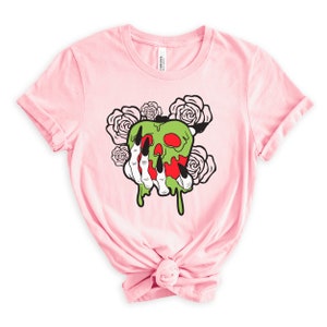 Pu&ograve; includere: Maglietta rosa con una mela verde e rossa con un viso di teschio e gocciolamenti di melma verde. Una mano scheletrica con lunghi artigli neri tiene la mela. Ci sono rose bianche intorno alla mela.