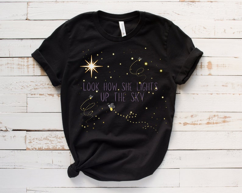 May include: Black t-shirt with a design of fireflies and stars. The text "Look how she lights up the sky" is printed on the shirt.