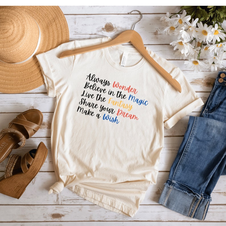 May include: A cream-colored t-shirt with a motivational quote printed on the front. The quote reads "Always Wonder Believe in the Magic Live the Fantasy Share your Dream Make a Wish". The text is in different colors, including red, orange, yellow, and blue.