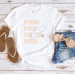 May include: White t-shirt with the text "Making Magic For The Mouse" in peach-colored lettering. The shirt is laid flat on a wooden surface with a pair of brown flip-flops and a pair of gold glitter mouse ears.