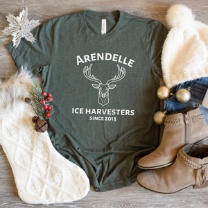 Może przedstawiać: Zielony T-shirt z białym nadrukiem jelenia z rogami i tekstem "Arendelle Ice Harvesters Since 2013". Koszulka jest otoczona przedmiotami o tematyce zimowej, w tym białą dzianinową skarpetą, białym puszystym kapeluszem i brązowymi zamszowymi butami.