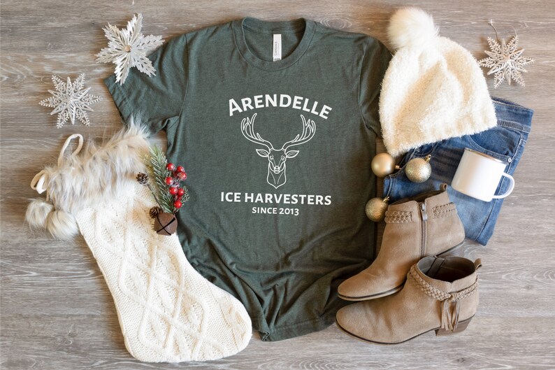 Może przedstawiać: Zielony T-shirt z białym nadrukiem jelenia z rogami i tekstem "Arendelle Ice Harvesters Since 2013". Koszulka jest otoczona przedmiotami o tematyce zimowej, w tym białą dzianinową skarpetą, białym puszystym kapeluszem i brązowymi zamszowymi butami.