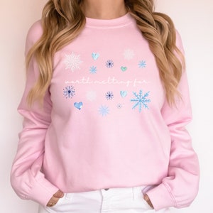 Puede incluir: Sudadera rosa con el texto "worth melting for" en letras blancas. La sudadera tiene un tema invernal con copos de nieve y corazones azules y blancos.