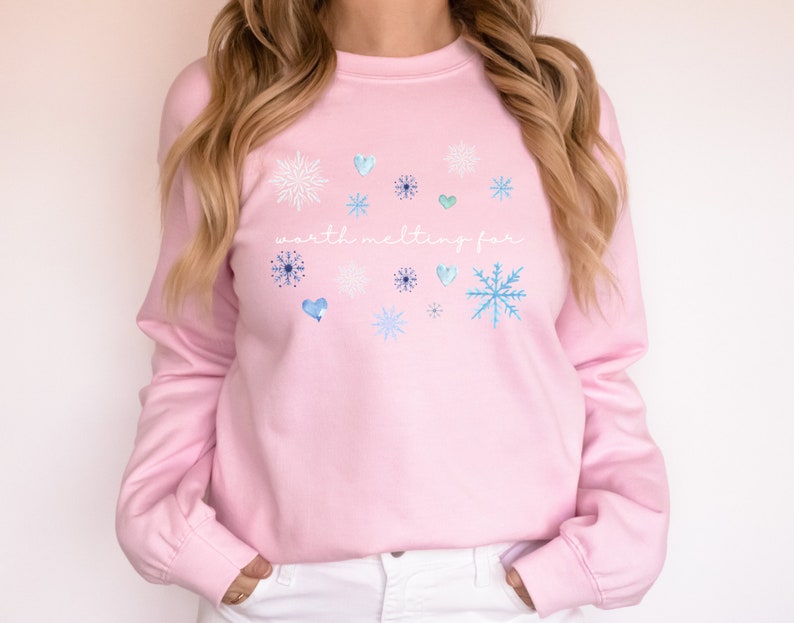 Puede incluir: Sudadera rosa con el texto "worth melting for" en letras blancas. La sudadera tiene un tema invernal con copos de nieve y corazones azules y blancos.