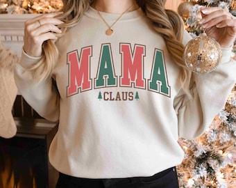 Op de afbeelding: Een crèmekleurig sweatshirt met de tekst "MAMA CLAUS" in rode, groene en blauwe letters met een kleine dennenboomgrafiek aan weerszijden van de tekst.