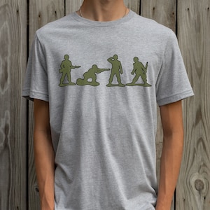 May include: A grey t-shirt featuring a graphic of five green toy soldiers in a line. The soldiers are in different poses, including standing, kneeling, and aiming a rifle.