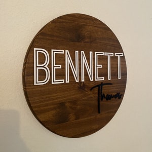 May include: A round wooden sign with the name "BENNETT" in white outlined letters and the name "Thomas" in black script underneath.