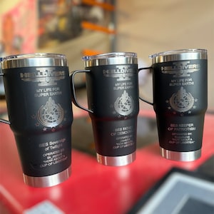 May include: Three black stainless steel travel mugs with a silver lid and handle. Each mug has a different design with the text "HELLDIVERS" and "MY LIFE FOR SUPER EARTH!" and a graphic of a rocket ship and a globe. The text "SES Sovereign of Twilight", "SES Father of Democracy", and "SES Keeper of Patriotism" is also on the mugs.