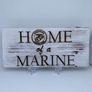 May include: A white wooden sign with the words "Home of a Marine" and the United States Marine Corps emblem.