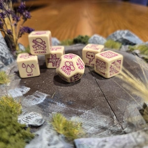 Puede incluir: Un conjunto de seis dados con diferentes símbolos y palabras, incluyendo "Hazard", "Wet", "Forest" y "Cave". Los dados son de color crema con letras rosas y están sobre una superficie marrón con un fondo texturizado gris y blanco.