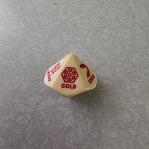 May include: Ivory-colored, multi-sided die with red text and symbols. The die features the word "COLD" and the words "THUNDER" and "FORCE". The die has a snowflake-like symbol and other geometric shapes. A unique game piece.