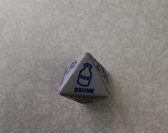 May include: A gray, triangular die with blue text and graphics. One side features a bottle graphic with the word "DRUNK" below it. Other sides have decorative patterns. The die is likely used in a drinking game.