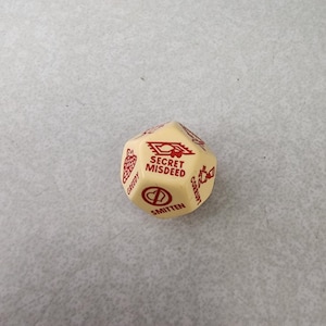 May include: A cream-colored, multi-sided die with red text and graphics. The die features words like "Greedy," "Smitten," and "Corrupt," along with a graphic of a heart and other symbols. The text "Secret Misdeed" is also visible.