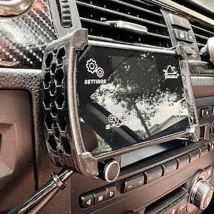 Pode incluir: Um dispositivo G-Force preto montado num carro, exibindo "SETTINGS" e "SCRAMBLE" no seu ecrã. O dispositivo é mantido no lugar por um suporte preto, com um design de favo de mel na lateral. O painel do carro é visível no fundo.