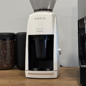 May include: A white Baratza coffee grinder with a black hopper and a black container. The grinder has a digital display and a dial on the side. A glass jar of coffee beans and a black container are to the left.