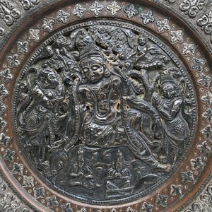May include: An antique metal plate with a detailed relief of a seated figure, other figures, and foliage. The plate has a dark, aged appearance and intricate patterns around the border. The plate's diameter is approximately 25 cm.