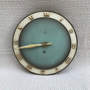 Può includere: Orologio rotondo vintage con quadrante azzurro e lancette e numeri romani color oro. Il quadrante dell'orologio ha la scritta "Junghans". L'orologio ha un bordo bianco screpolato e una cornice in metallo dorato.