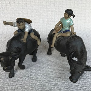 May include: Two black water buffalo figurines, each with a small child rider. The children wear light-colored shirts and shorts, and hold hats. The buffalo have curved horns and are posed on a neutral background.