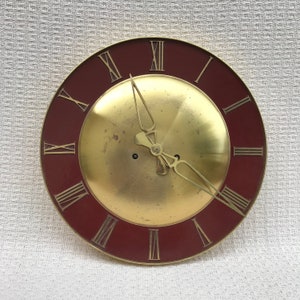 Op de afbeelding: Een gouden wijzerplaat met Romeinse cijfers en een bordeauxrode buitenring. De wijzers van de klok staan ​​op 10:10.