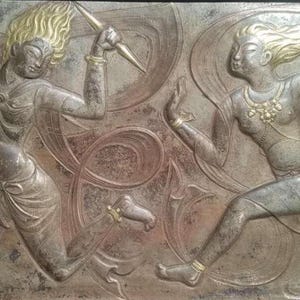 May include: A detailed relief sculpture showing two figures in action. One figure holds weapons, the other appears to be dancing. The figures have gold-coloured hair and accents, set against a muted, metallic background.