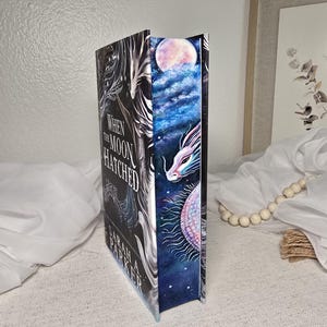 May include: A hardcover book titled When the Moon Hatched by Sarah A. Parker. The front cover is black with white text, while the page edges feature a detailed painting of a white dragon, a glowing moon, and a starry blue night sky.