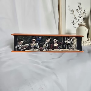 May include: A painted wooden box with a scene of people sitting at a table with wine glasses and a plate of fruit. The box is painted in a dark, realistic style with a brown background and orange trim.