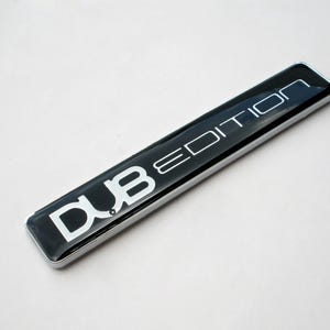 May include: A rectangular black emblem with the words "DUB EDITION" in white, outlined lettering. The emblem has a shiny, reflective surface and a silver border. The item is likely an automotive accessory.