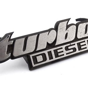 Może przedstawiać: Srebrny i czarny emblemat "Turbo Diesel". Słowo "turbo" jest w stylizowanej czcionce nad słowem "diesel". Emblemat ma lekko zwietrzały wygląd, co sugeruje, że jest to starsza część.