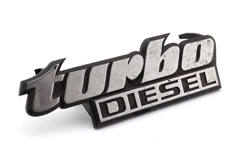 Może przedstawiać: Srebrny i czarny emblemat "Turbo Diesel". Słowo "turbo" jest w stylizowanej czcionce nad słowem "diesel". Emblemat ma lekko zwietrzały wygląd, co sugeruje, że jest to starsza część.